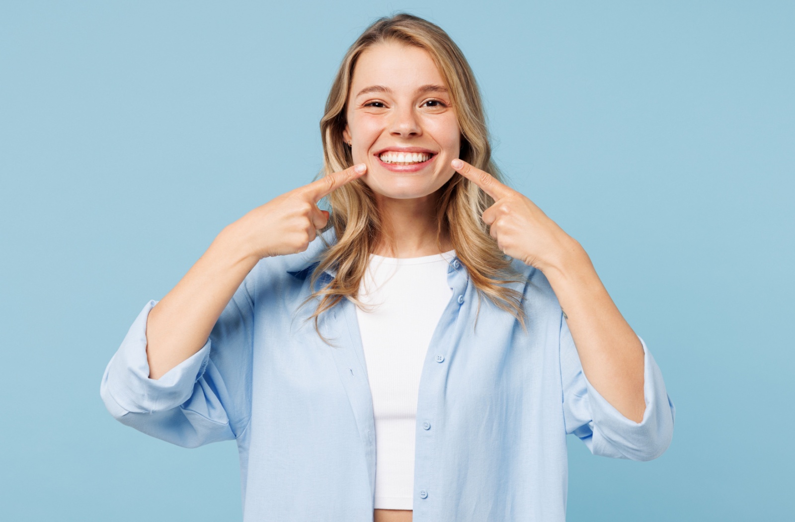 A young adult uses both pointer fingers to point to their bright, white smile while they stand in front of a blue background.